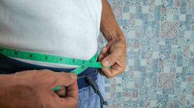 Yakın çekim, belinin çevresini yeşil bir mezura ile ölçen bir kişiyi içerir. Kişi beyaz gömlek ve mavi pantolon giyiyor. Arka plan desenli bir duvar. Görüntü sağlık, zindelik ve ağırlık kavramını aktarır.