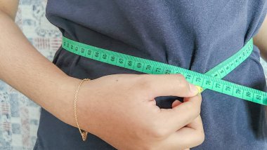 Yakından çekilen bir fotoğraf, kişinin belini ölçmek için yeşil bir mezura kullanır. Kişi koyu mavi bir tişört giyiyor, ve kolları ve elleri görünür, ölçüm bandını tutuyor. Odak noktamız ölçüm süreci..