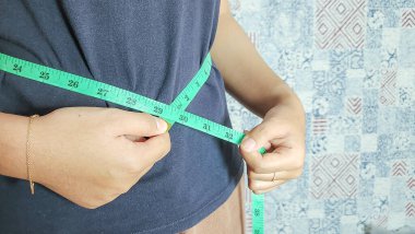 Bir bireye yeşil bir mezura kullanılarak bel çevresini ölçtüğü gösterilir. Cinsiyeti açıkça belli olmayan kişi koyu mavi bir bluz ve kahverengi pantolon giyiyor. Arka planda, nötr bir ayar oluşturan desenli bir duvar var..