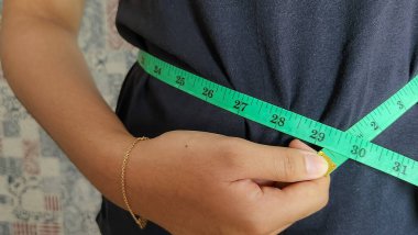 Bir kişi yeşil bir mezura kullanarak bel ölçüsünü ölçerken görülüyor. Görüntü, vücut ölçüm sürecine odaklanır, muhtemelen sağlık, fitness veya ağırlık yönetimi ile ilgilidir. 