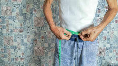 Beyaz tişört ve mavi şort giyen bir adam, yeşil bir mezura ile belini ölçüyor. Görüntü kilo verme ve vücut ölçümleri kavramını vurguluyor. Arkaplanda desenli bir duvar var.. 