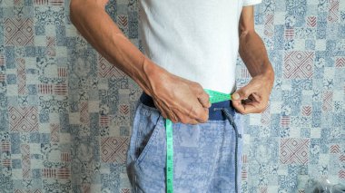 Bir adam, beyaz tişört ve mavi çizgili pantolon giyerken belini yeşil bir şeritle ölçüyor. Görüntü sağlık ve zindelik üzerine odaklanarak, kilo kaybı veya sağlık izleme potansiyelini gösteriyor. Arkaplanda desenli bir duvar var.. 