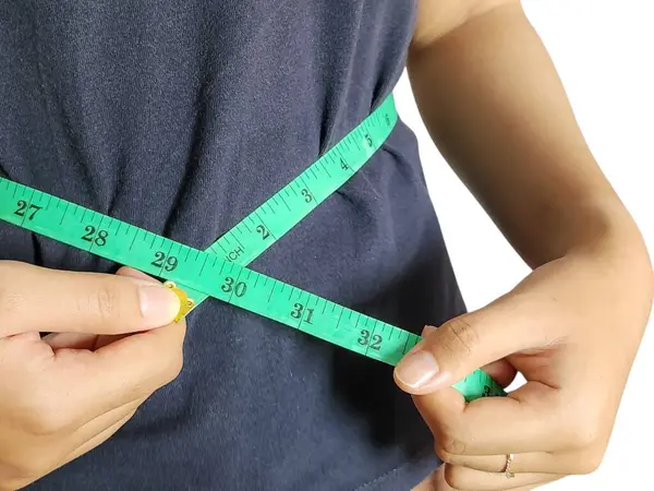 Bir kişi, muhtemelen bir kadın, yeşil bir mezura ile belini ölçerken görülüyor. Görüntü vücut ölçüleri alma, sağlık ve uygunluğu vurgulama eylemine odaklanıyor. Koyu renkli bir bluz giyiyor, ve arka planı düz beyaz..