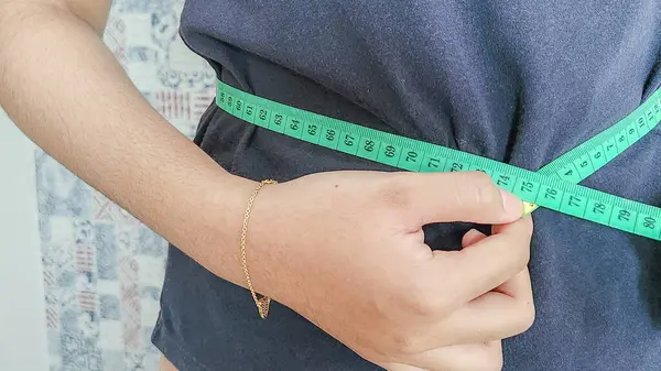 Bel çevresini yeşil bir mezura ile ölçen bir insan, vücut şekli, sağlık ve zindelik kavramını vurguluyor. Görüntü, ölçüm bandını tutan kişinin eliyle bel bölgesine odaklanıyor..
