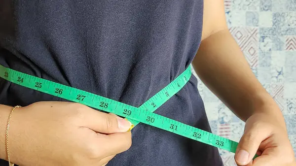 Yakından bakınca, lacivert bir gömlek giyen birinin yeşil bir mezura ile belini ölçtüğünü görüyoruz. Mezura belin etrafına sarılmış, ve kişi onu elleriyle tutuyor. Odak noktamız ölçüm süreci..