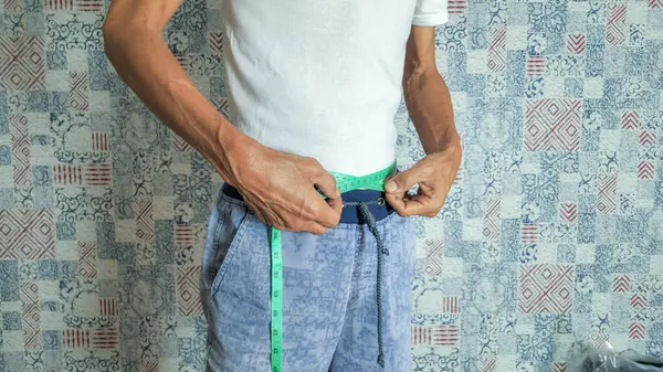 Evde yaşlı bir adam gösteriliyor, yeşil bir mezura ile belini ölçüyor. Beyaz tişört ve askılı mavi pantolon giyiyor. Arka planda mavi, beyaz ve kırmızı tonlarda desenli bir duvar kağıdı yer almaktadır..