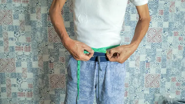 Yakın plan çekimde, yeşil bir mezura ile belini ölçen bir adam görülüyor. O, beyaz bir tişört ve mavi bir şort giyiyor. Arka plan, tekrarlayan mavi ve kırmızı geometrik şekilli dokulu bir duvar..