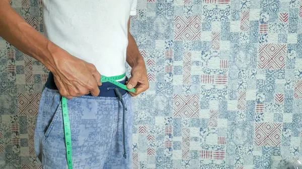 Bir adam, muhtemelen orta yaşlı ya da daha yaşlı, yeşil bir mezura ile belini ölçüyor. O, beyaz bir tişört ve asma ipli mavi desenli pantolon giyiyor. Arka planda mavi ve kırmızı tonlarında yama tasarımlı dekoratif bir duvar var.. 