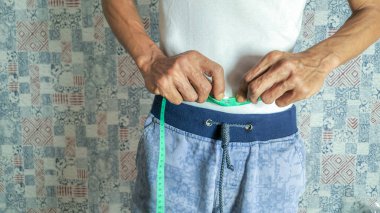 Bir kişinin yeşil bir mezura kullanarak belini ölçtüğü görülüyor. Beyaz gömlek ve mavi pantolon giyiyorlar. Arka planda geometrik ve çiçekli tasarımların karışımından oluşan desenli bir duvar var..
