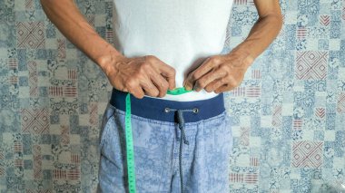 Yeşil bir mezura ile belini ölçen bir adamın yakın çekim görüntüsü. O, beyaz bir tişört ve mavi bir şort giyiyor. Arka plan desenli bir duvar. Bu görüntü sağlık, zindelik, kilo kaybı ve vücut ölçüm kavramları için uygundur..