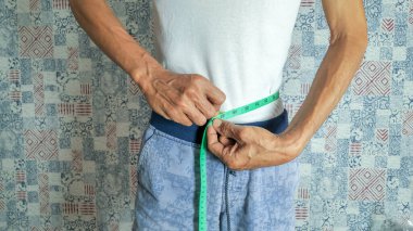 Bir adam, arka planda yeşil bir mezura ile belini ölçerken görülüyor. Beyaz bir atlet ve mavi bir şort giyiyor. Görüntü, sağlık, kilo kaybı ve vücut ölçümü temalarını iletir. Odak noktası ölçüm eylemi