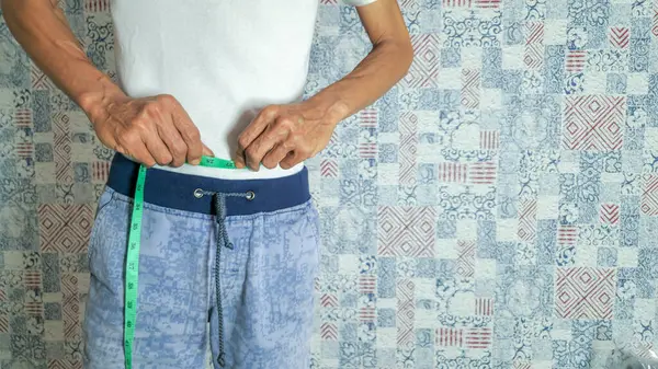 Bir adama yeşil bir mezura ile belini ölçtüğü gösterilir. Beyaz bir gömlek ve mavi bir şort giyiyor. Arka planda kapalı bir yerde duruyor. Görüntü ağırlık yönetimi, sağlıklı yaşam tarzı ve vücut ölçümleri kavramını yakalıyor..