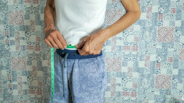 Orta yaşlı bir adam yeşil bir mezura ile belini ölçer. Beyaz bir atlet ve mavi bir şort giyiyor. Arka plan desenli bir duvar. Görüntü, sağlık, kilo kaybı ve vücut ölçümlerine odaklanılmasını öneriyor..