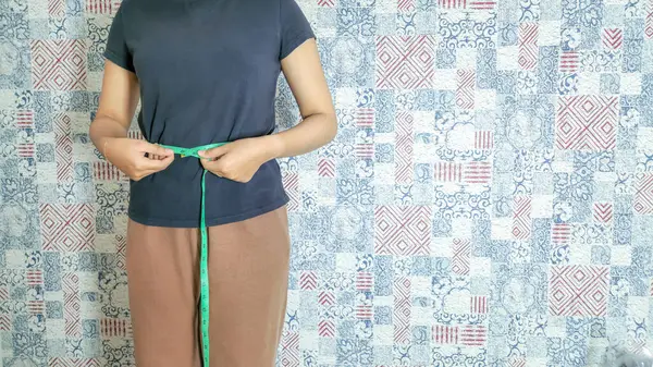 Koyu mavi tişörtlü ve kahverengi etekli bir kadın yeşil mezura kullanarak belini ölçüyor. O, desenli bir duvar kağıdının önünde duruyor. Görüntü sağlık, zindelik ve vücut ölçümlerine odaklanıyor, kilo kaybı ile ilgili makaleler için ideal.