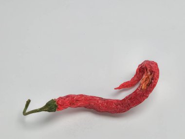 Yakın plan bir stüdyoda, parlak beyaz bir arkaplanda tek bir kırmızı kırmızı biber sergilenir. Biber buruşuk bir doku ve yeşil sapla kıvrılmış ve susuz kalmış halini vurguluyor. Işıklandırma biberin şeklini ve rengini vurguluyor.