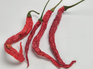 Beyaz bir yüzeye dizilmiş dört kurutulmuş kırmızı biberli yakın plan fotoğraf. Biberler buruşuk ve koyu kırmızı bir renge sahip, yeşil sapları var. Biberlerin dokularını ve şekillerini vurguluyor..
