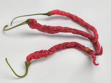 Temiz beyaz bir yüzeye üç kurutulmuş kırmızı biber yerleştirildi. Biberler buruşmuş ve kurumuş durumlarını gösteren yeşil sapları var. Görüntü, biberlerin dokusunu ve rengini vurgulayan yakın plan bir fotoğraf..