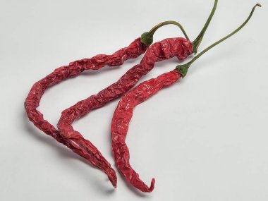 Bu resimde, temiz beyaz arka planda sıralanmış üç kurutulmuş kırmızı biber sergileniyor. Biberler kırışık bir doku ve yeşil saplı canlı kırmızı bir renk sergiler. Biberlerin doğal biçimini ve rengini vurgulayan kompozisyon basittir..