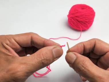 İğne ve pembe iplik tutan ellerin yakın plan çekimi. Bir el ipliği iğnenin deliğinden geçirir. Arka planda bir pembe iplik topu görülebilir. Görüntü beyaz arkaplana karşı iyi ışıklandırılmış. .