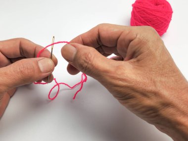 Yakın plan çekimde, beyaz bir arka planda parlak pembe iplikle bir iğneyi dikkatlice iplikçiklerken görülüyor. İğne altın renginde ve iplik parlak, göz alıcı bir pembe. Görüntü, ellere ve dikiş sürecine odaklanıyor..