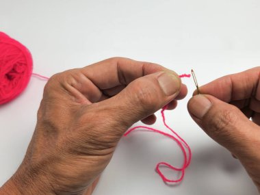 Yakın plan çekimde elleri, parlak pembe iplikle iğneyi dikkatlice iplikçikliyor. Resim, bir insanın ellerini bir dikiş projesinde çalışırken gösteren titiz bir dikiş veya el işi sürecini vurguluyor. Beyaz arka plan temiz bir arkaplan sağlar..