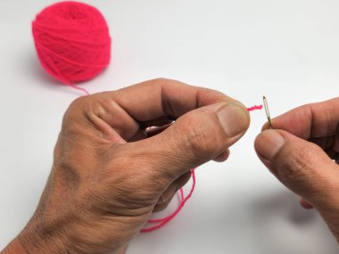 Yakından çekilen bir fotoğraf, elleri dikkatle bir iğnenin gözüne pembe iplik sokuyor. Görüntü, ellerin detaylarını, ipliği ve iğneyi temiz beyaz arka planda yakalar. Pembe bir iplik yumağı odaklanamaz.