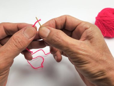 Yakın plan çekimde, beyaz arka planda parlak pembe iplikle altın bir iğneyi dikkatlice iplikçiklerken görülüyor. Dikiş ya da zanaatın titiz eylemine odaklanılıyor. Arka planda iplik yumağı görülüyor...