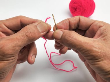 Bir insanın ellerinin pembe bir ipliği iğnenin deliğinden geçirdiği yakın plan bir fotoğraf. Eller iğneyi ve ipliği beyaz arka planda tutuyor. Arka planda bir pembe iplik topu görülebilir. Görüntü odak duygusunu iletir.