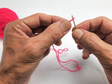 Altın dikişli iğnenin gözüne pembe iplik bağlayan ellerin yakın plan çekimi. Resim beyaz bir arkaplana doğru parlak bir şekilde aydınlatılıyor, el dokusunun ve ipliğin renginin detaylarını gösteriyor. Arka planda pembe iplik yumağı oturuyor..