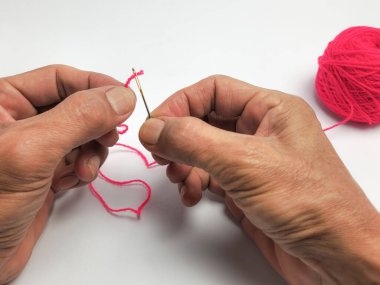 Tepeden çekilen bir fotoğraf, pembe iplikle iğne ipliği tutan elleri yakalıyor. Görünür deri dokusu ve yaşı olan eller iğneyi ve ipliği dikkatlice manipüle eder. Arka planda pembe bir iplik yumağı duruyor ve sahne beyaz bir ipliğe karşı hazırlanıyor..