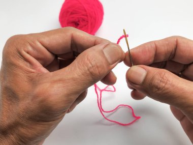 Yakın çekim, bir insanın ellerini parlak pembe iplikle iğneyi dikkatlice iplikçiklerken gösterir. Elleri yaşlanmış. Tecrübe olduğunu gösteriyor. Beyaz arkaplan iplik ve iğneyi vurgular, basit ve temiz bir kompozisyon oluşturur..