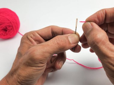 Yakın çekim, parlak pembe iplikle iğne ipliğini dikkatlice yakalar. Resim, ellerin maharetini ve ilgili araçları gösteren, dikiş işleminin ayrıntılı sürecine odaklanır. Arka planda pembe iplik yumağı oturuyor..