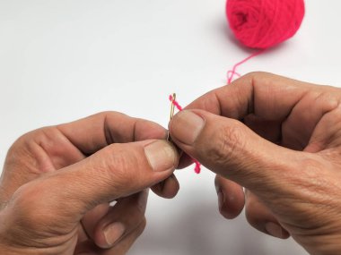 Yakın plan çekimde, parlak pembe iplikle bir iğneyi dikkatlice iplikçikleyerek. Görüntü, zanaat için gereken detayı ve odağı vurgulayan titiz el dikme sürecini gösteriyor. Arka plan bembeyaz, elleri vurguluyor..