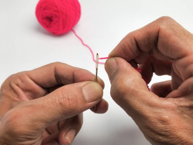Bir insanın ellerinin pembe bir ipliği iğnenin deliğinden geçirdiği yakın plan bir fotoğraf. Pembe iplik yumağı arka planda, temiz beyaz arka planda duruyor. Görüntü, ellerin ve ipliğin detaylarını vurgular, etkinliğe odaklanır..