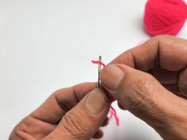 Bir insanın ellerinin pembe iplikle iğneye iplik geçirmesini yakından çek. Eller ipliği iğnenin gözünden geçirmek için dikkatlice yönlendiriyor. Pembe iplik yumağı bulanık arkaplanda görünür..