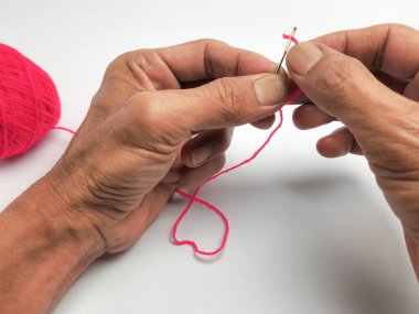 Yakından çekilen bir fotoğraf, düz beyaz bir arkaplana uzanan parlak pembe iplikle bir iğneyi dikkatlice yakalar. Görüntü, ellerin, iğnenin ve ipliğin detaylarını vurgular, el sanatları sürecini gösterir..