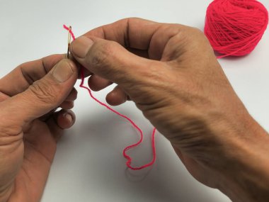 Bir insanın ellerinin parlak kırmızı iplikle iğneye iplik geçirmesini yakından çek. Arka planda bir kırmızı iplik yumağı oturuyor. Görüntü, dikiş, el işi ve ilgili araçları vurguluyor. Ellere ve iğneye odaklanmış durumda..