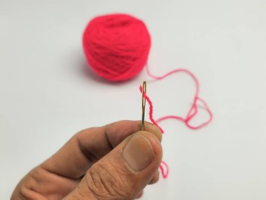 Bu resim, parlak pembe bir iplik yumağının yanında pembe iplikle dikilmiş bir dikiş iğnesini tutan bir eli gösteriyor. Kompozisyon, temiz beyaz bir arkaplan karşısında dikiş konseptini vurguluyor. Dikkatimizi iğnenin detaylarına vermeliyiz..