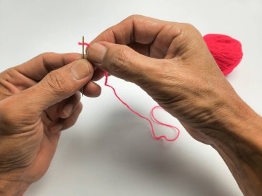 Bir insanın ellerinin parlak pembe bir iplikle düz beyaz bir arka plana iplik geçirdiği yakın plan bir fotoğraf. Resim dikiş sürecini vurgular, iğne ve iplik merkezi odak noktasıdır.