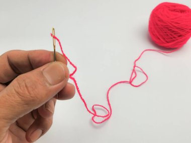 Yakın çekim, temiz beyaz bir arkaplana karşı parlak pembe iplikle dikiş iğnesini iplikçikleyen bir eli gösteriyor. Görüntü iğneyi, ipliği ve onu tutan eli vurgular, arka planda görünen bir iplik yumağıyla.. 