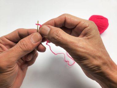 Bir insanın ellerinin parlak pembe bir iplikle temiz beyaz bir arkaplana iplik geçirdiği yakın plan görüntüsü. Görüntü, iğnenin ve ipliğin detaylarını vurguluyor. Ellerin malzemeleri dikkatlice manipüle ettiğini gösteriyor..