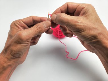 Tepeden çekilen yakın çekim, bir insanın ellerini parlak pembe iplikle iğneyi dikkatlice ipliklerken gösteriyor. Yaşını gösteren eller iğneyi ve ipliği temiz beyaz bir arkaplanda tutuyor. Arka planda küçük bir pembe iplik topu görünür..