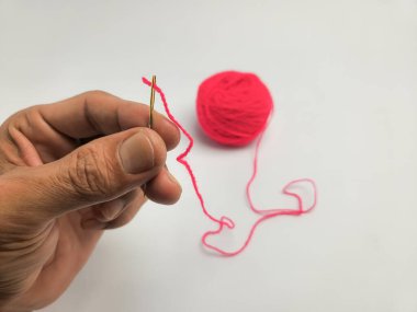 Yakından çekilen bir resimde parlak kırmızı iplikle işlenmiş dikiş iğnesini tutan bir el görülüyor. İğne çerçeve üst yakın konumdadır, iplik arka planda bir iplik topu doğru uzanır.