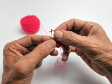 Yakından çekilen fotoğraf, ellerini dikkatlice pembe iplikle iğne ipliğine sokuyor. Arka planda bir pembe iplik topu görülebilir. Resim beyaz bir yüzeyde güzel bir ışıkla çekiliyor, ellerin ve ipliğin detaylarını vurguluyor.. 