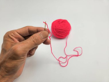 Yakından çekilen bir el, parlak kırmızı iplikle bir iğneyi dikkatlice yakalıyor. İğne parmaklar arasında tutulur ve iplik gözün içinden çekilir. Temiz beyaz arka planda kırmızı bir iplik yumağı duruyor..