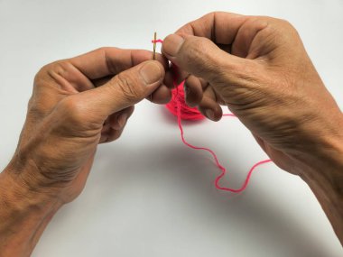 Yakın çekim, bir insanın ellerini pembe iplikle iğne ipliğine bağlarken gösteriyor. Elleri yaşlanmış, bu da yaşlı bir yetişkin olduğunu gösteriyor. Resim beyaz bir arkaplanda çekilir, temiz ve basit bir kompozisyon oluşturur.