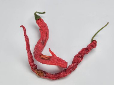 Üç kurutulmuş kırmızı biberin yer aldığı bir makro çekim, buruşuk dokularını ve parlak renklerini temiz beyaz arka plana karşı sergiliyor. Biberler ilginç bir kompozisyon oluşturmak için düzenlenmiş..