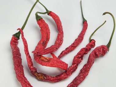 Yakın çekim, yüksek açı çekim vitrinleri. Beyaz arka planda beş kurutulmuş kırmızı biber. Biberler kırış kırış ve koyu kırmızı saplı, yeşil saplı. Resim biberlerin dokusunu ve detaylarını vurgular.