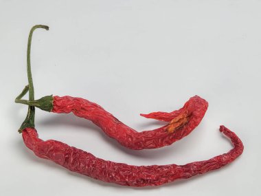 Yakın çekim bir stüdyo çekiminde iki kurutulmuş kırmızı biber, temiz beyaz bir arkaplana karşı. Biberler, kurumuş sürecin göstergesi olarak buruşuk bir doku ve zengin bir renk sergiliyor. Bileşim, biberin benzersiz şekillerini ve biçimlerini vurgular..