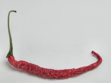 Tek bir kurutulmuş kırmızı biber, temiz beyaz arka planda izole edilir. Biber uzatılmış ve kıvrımlıdır, desenli derisini ve yeşil sapını gösterir. Görüntü iyi aydınlatılmış, biberin rengini ve şeklini vurguluyor..
